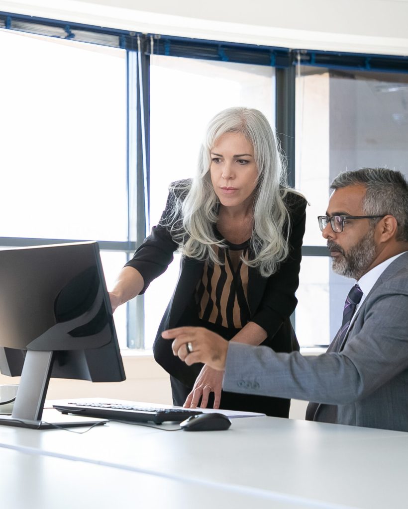 Serious colleagues watching and discussing content on computer monitor, pointing at display and talking while sitting in meeting room with panoramic window. Business communication concept